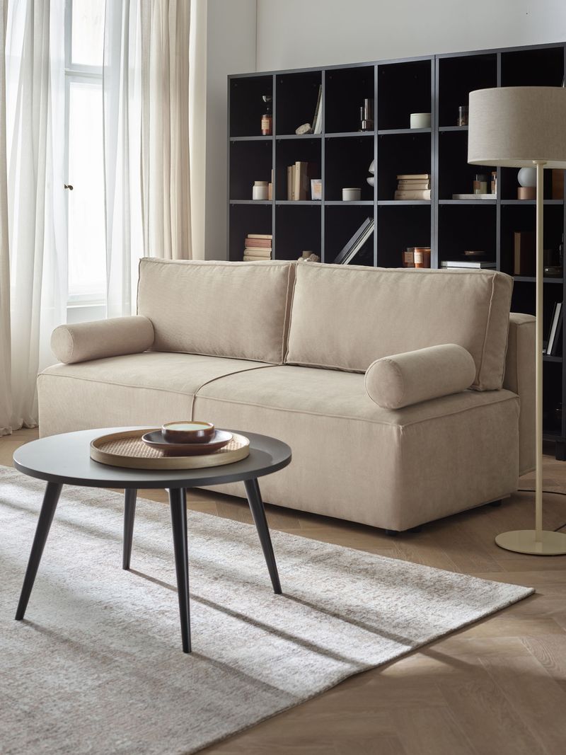 Jasna sofa Solis stoi przed ciemnym regałem