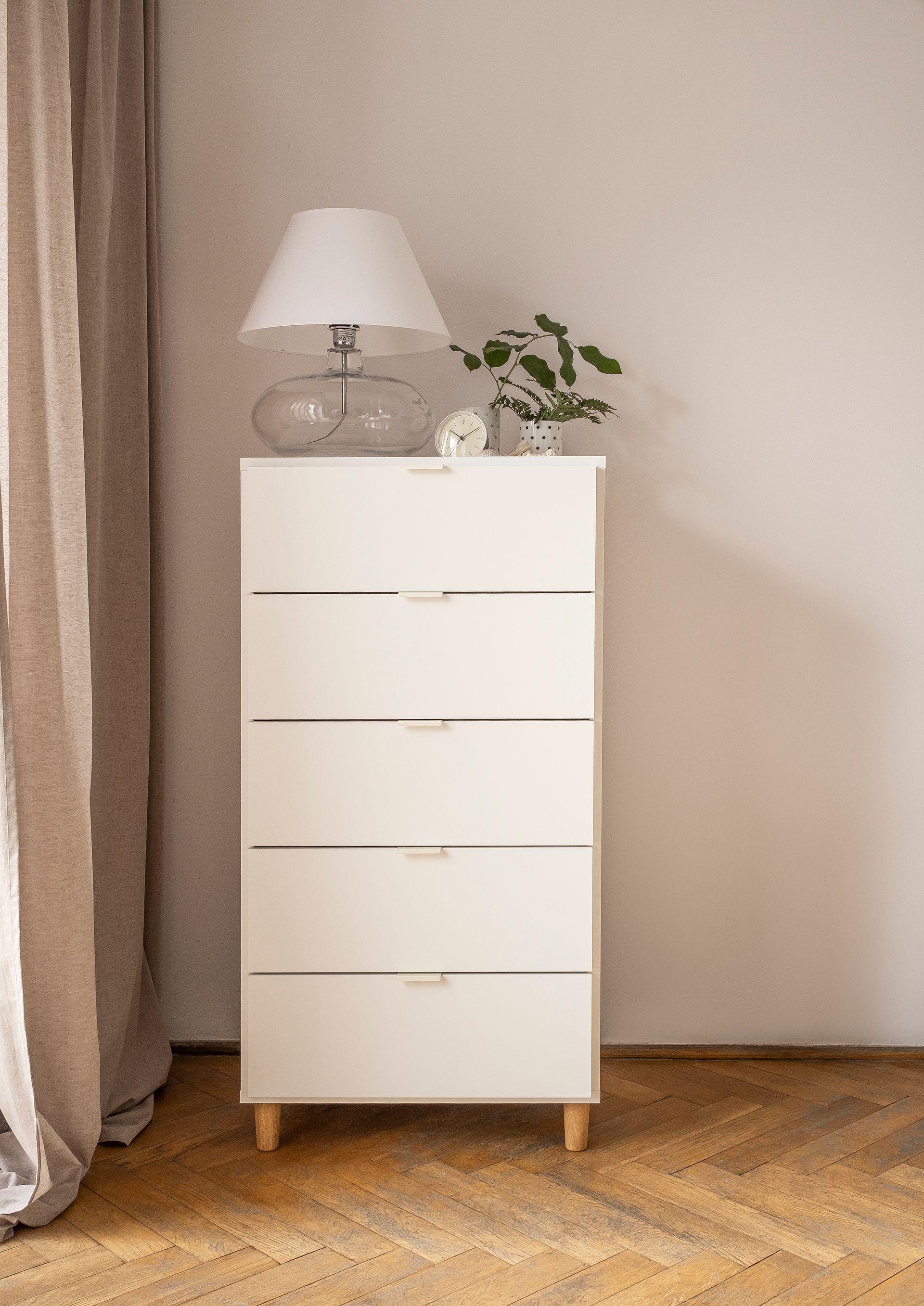 Wysoka biała komoda z lampą i roślinami na jasnej ścianie.