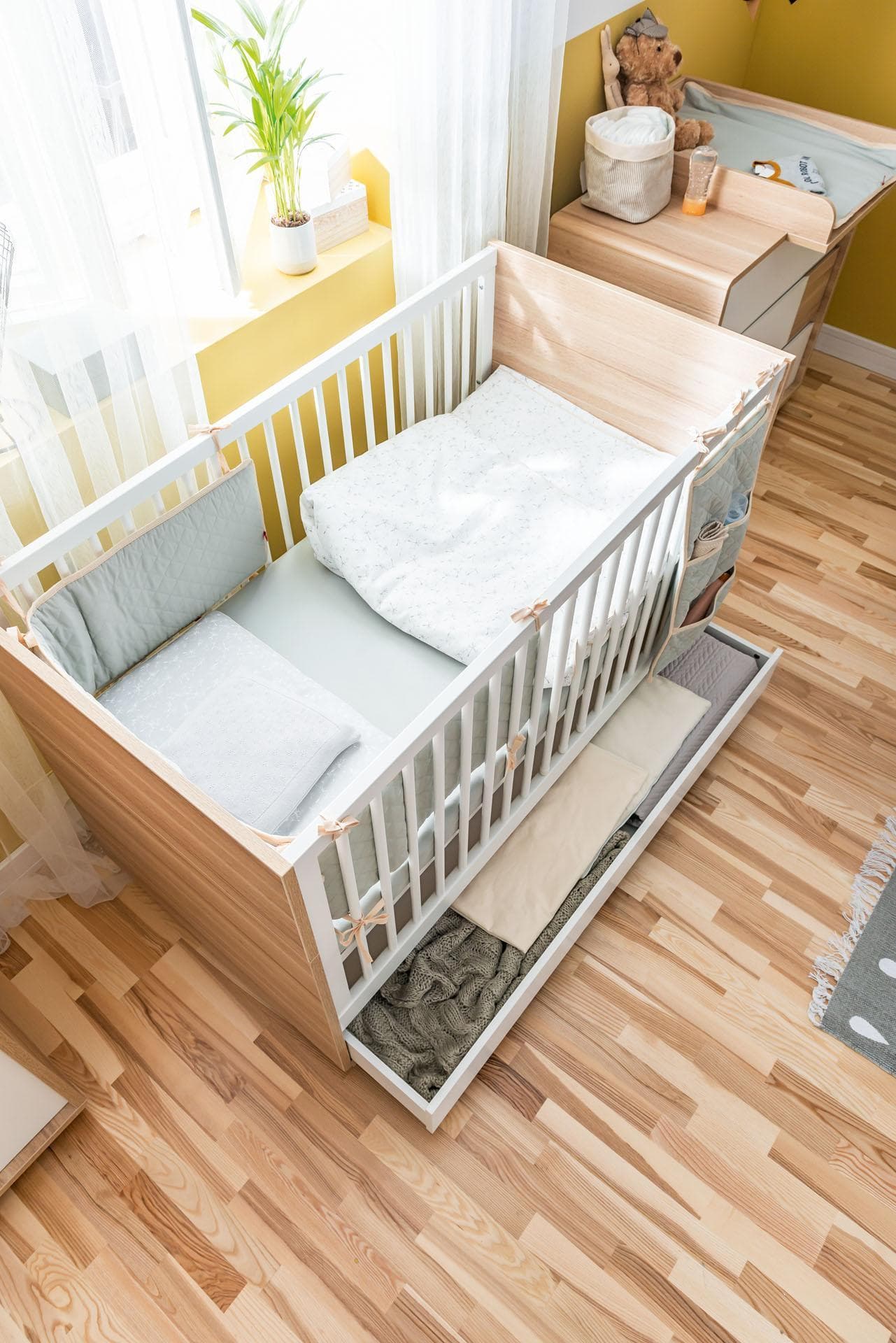 Łóżeczko z wysuwaną szufladą na pościel, utrzymane w jasnej i ciepłej stylistyce.