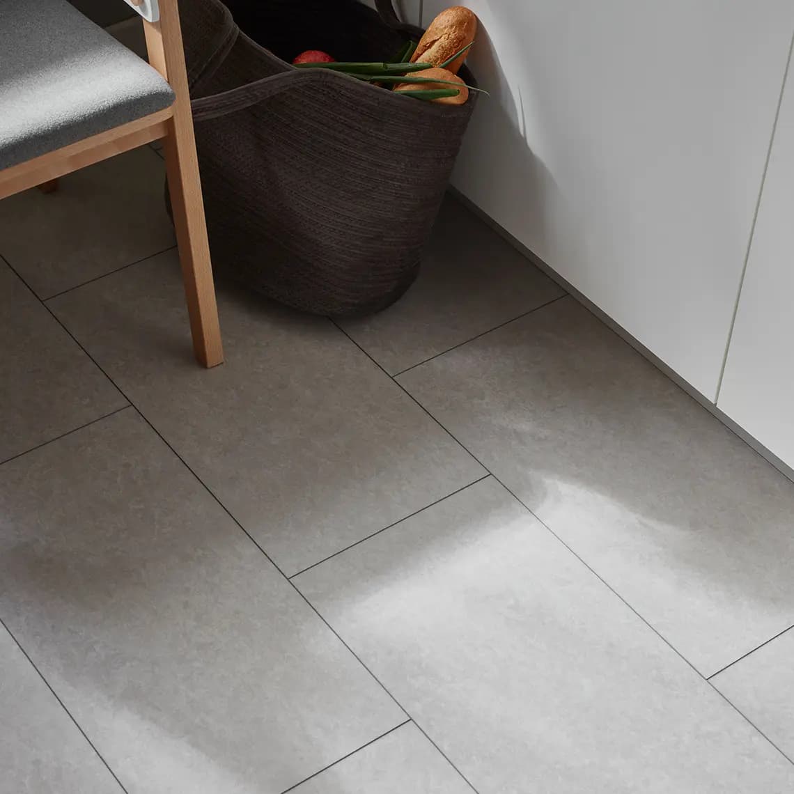 Zbliżenie na szarą podłogę imitującą beton w kuchni, obok drewnianego krzesła i kosza na zakupy.